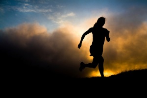 A woman runs on a trail silhouetted by a setting sun shrouded by fog.